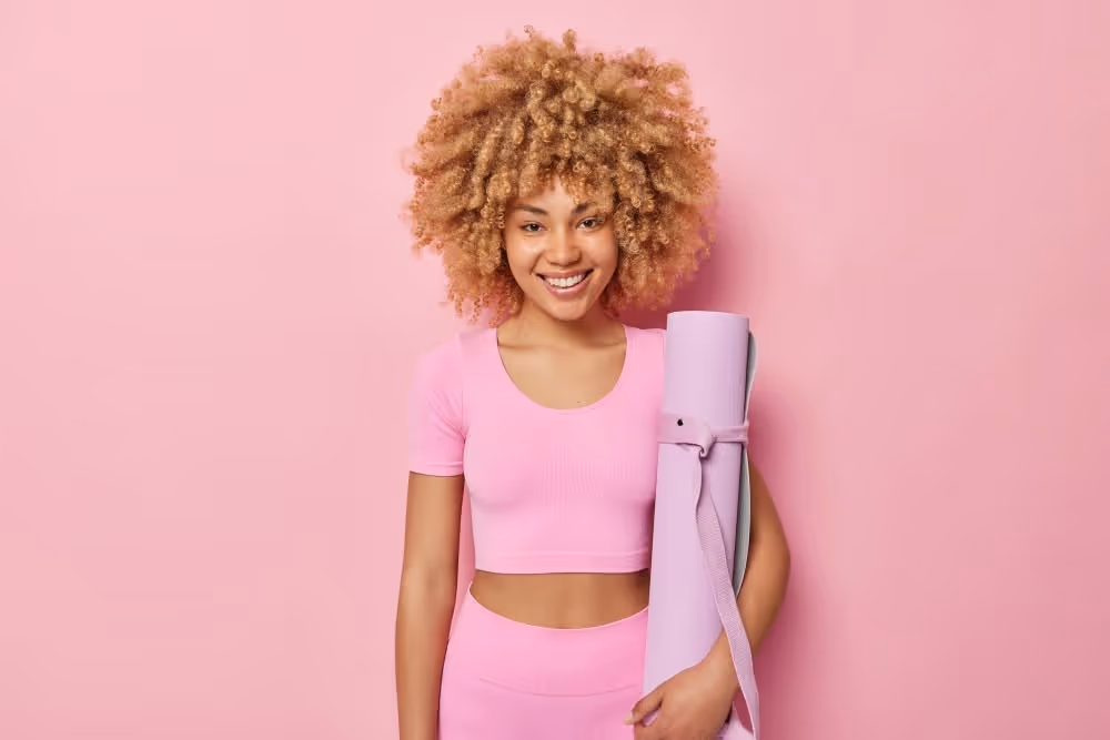 Smiling woman with curly hair wearing pink athletic wear holding a rolled-up yoga mat against a pink background.