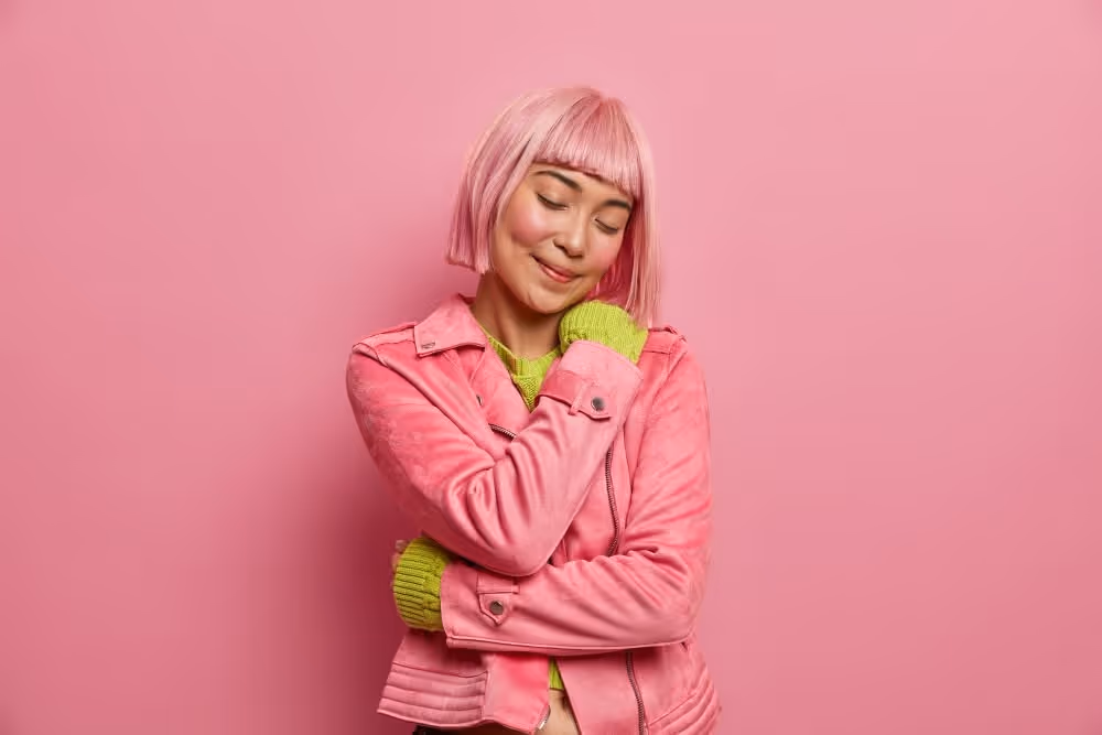 Smiling woman with pink bob haircut wearing a pink jacket and green sweater against a pink background.
