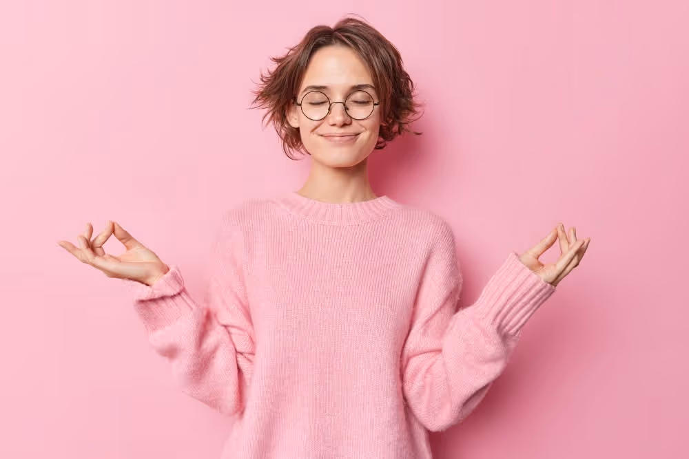 Young woman with short hair wearing round glasses and a pink sweater meditating with eyes closed and hands in a mudra gesture against a pink background.