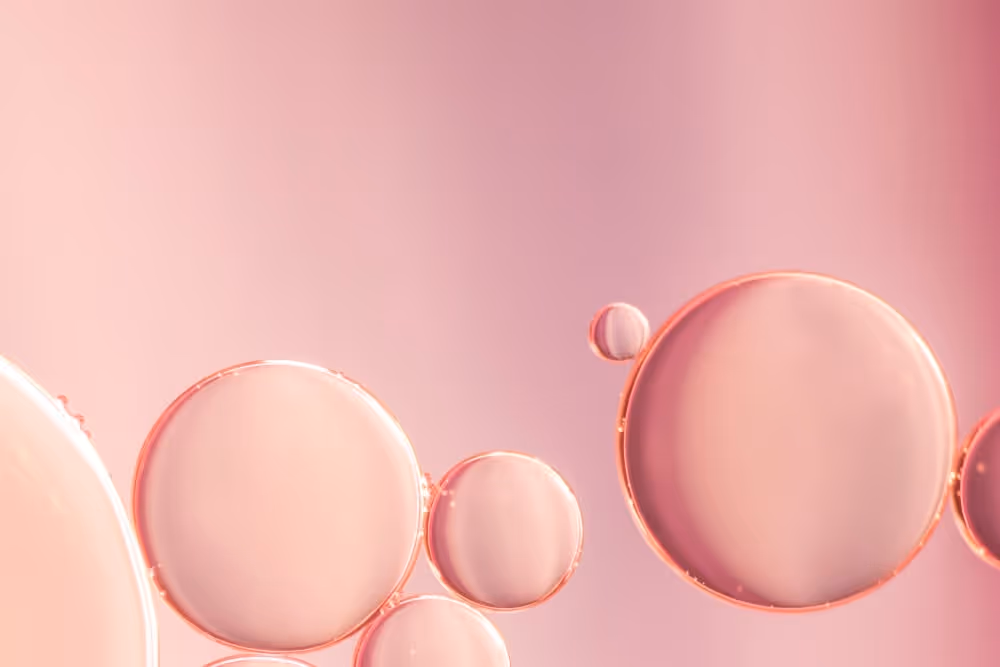 Close-up of translucent pink oil droplets suspended in liquid with a soft pink background.