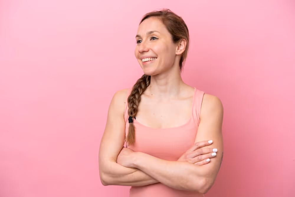 Smiling woman with braided hair wearing a pink tank top standing against a pink background with arms crossed.