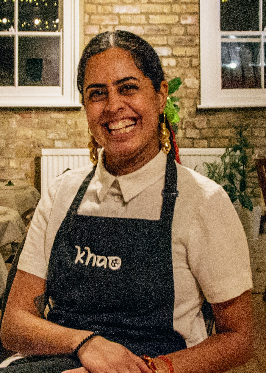 Smiling woman wearing a white shirt and a black apron with the logo 'kha' in a warmly lit room with brick walls and windows.