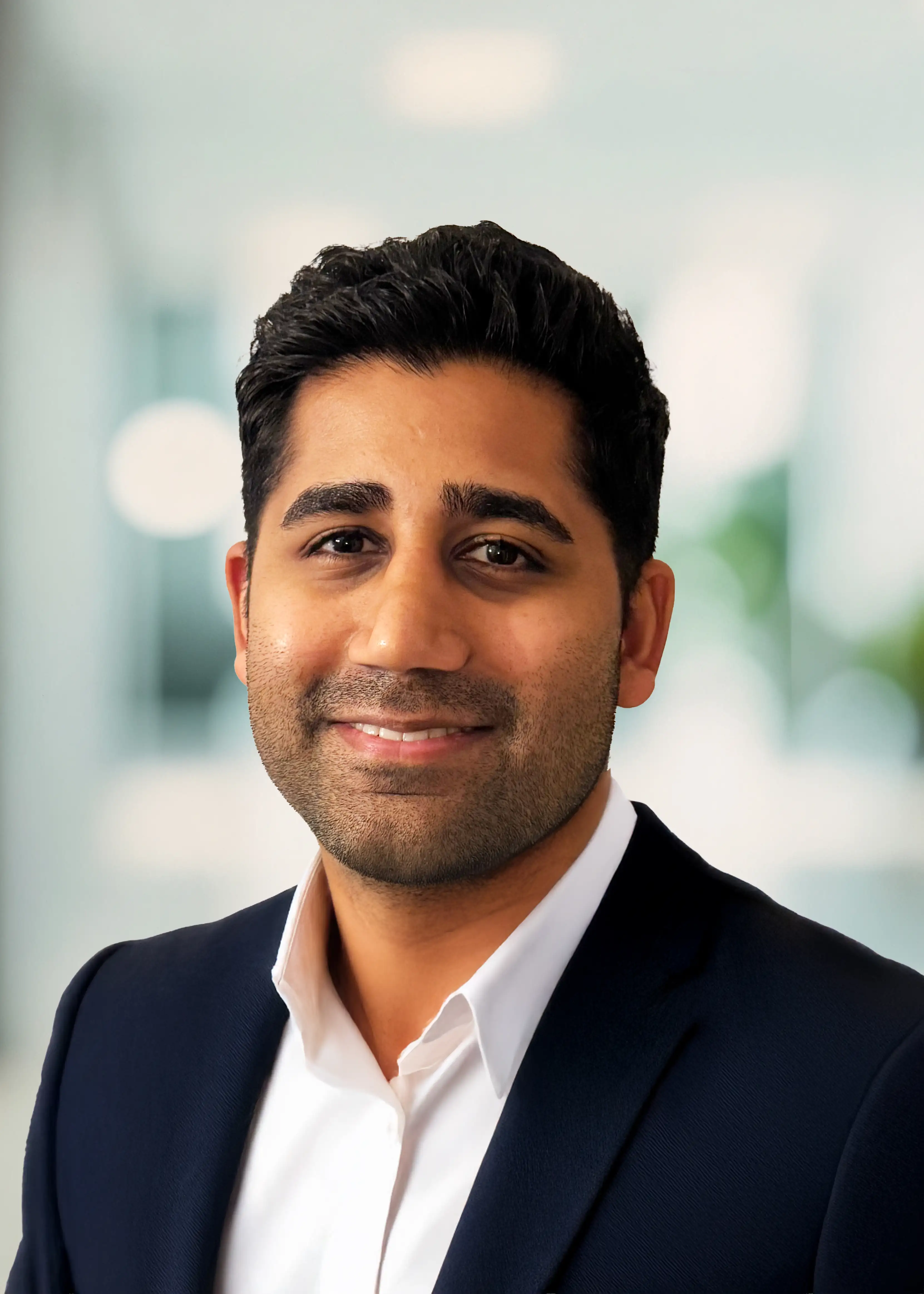 Smiling man with dark hair and beard wearing a white shirt and dark blazer in an office setting.