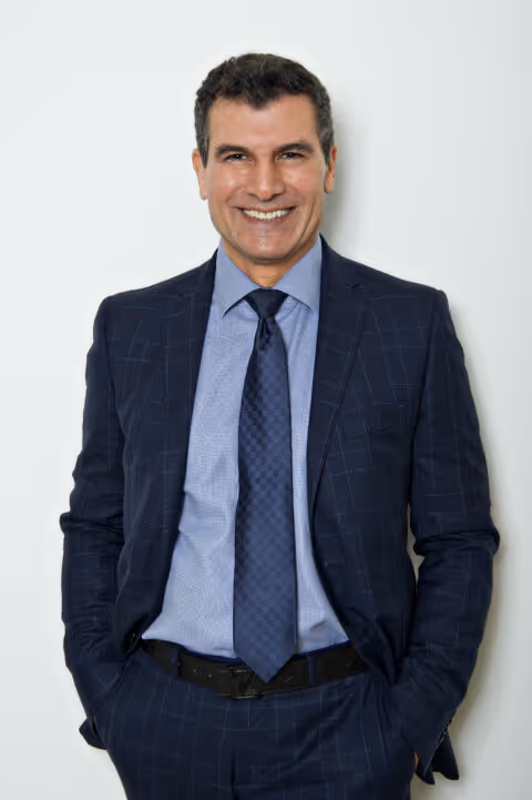 A man in a dark suit and tie stands smiling with hands in his pockets.