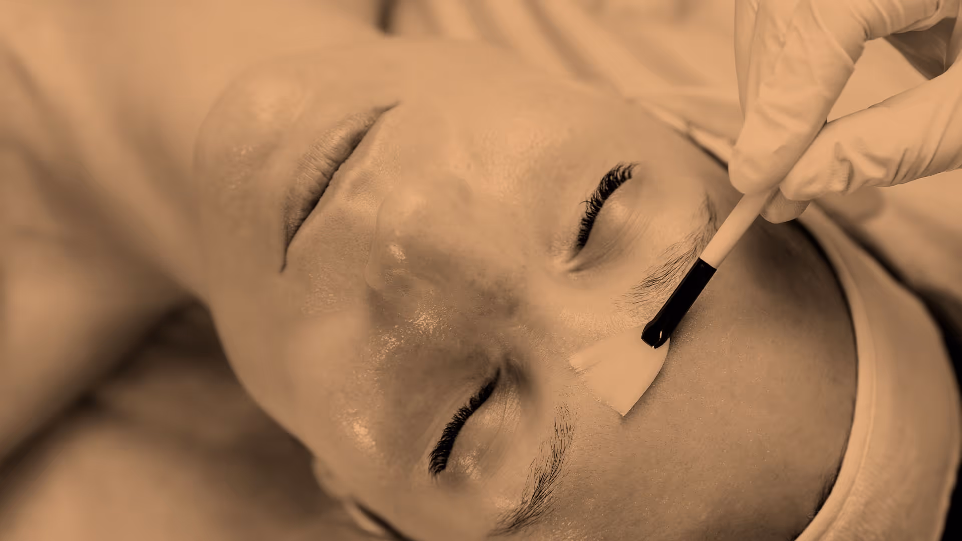 A person receives a facial treatment as a gloved hand applies a brush to their forehead.