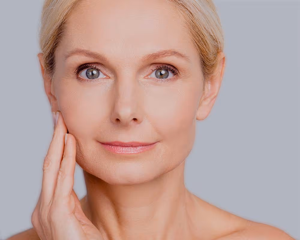 A woman with short blonde hair touches her face while looking directly at the camera.