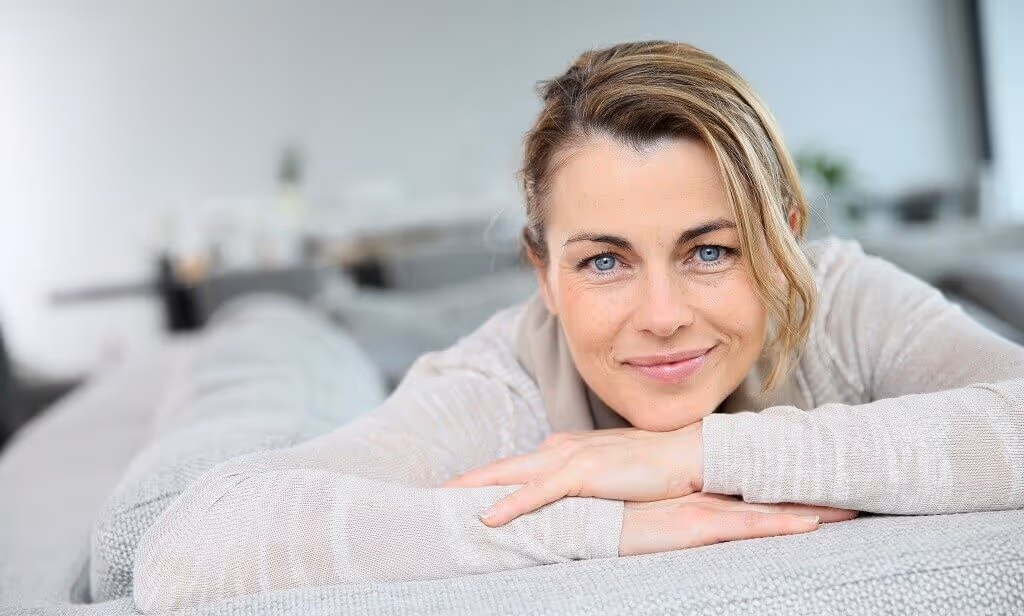 A woman with blonde hair is lying on a couch, smiling and resting her chin on her hands.