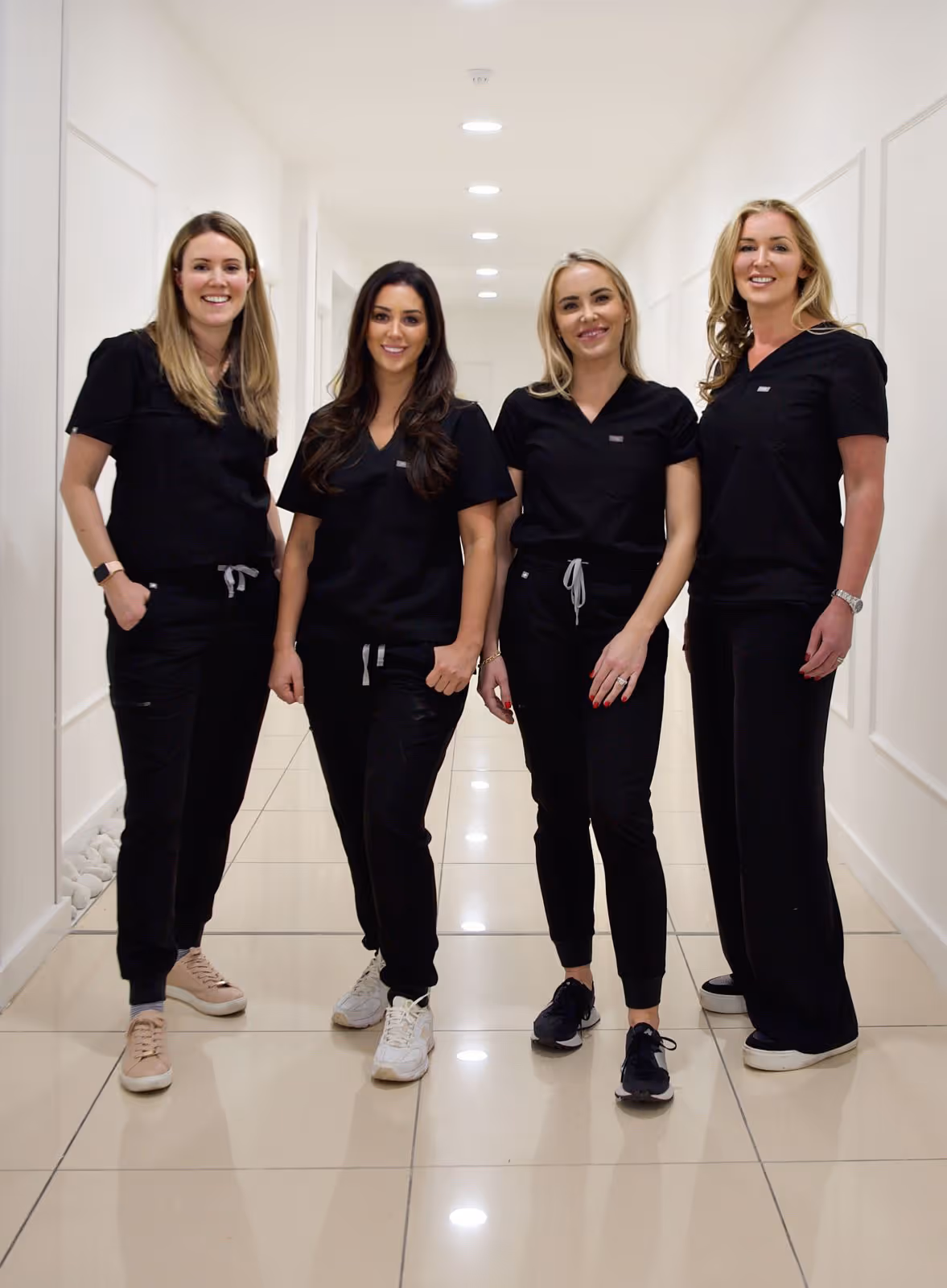Four women standing in a hallway, wearing black scrubs, smiling at the camera.
