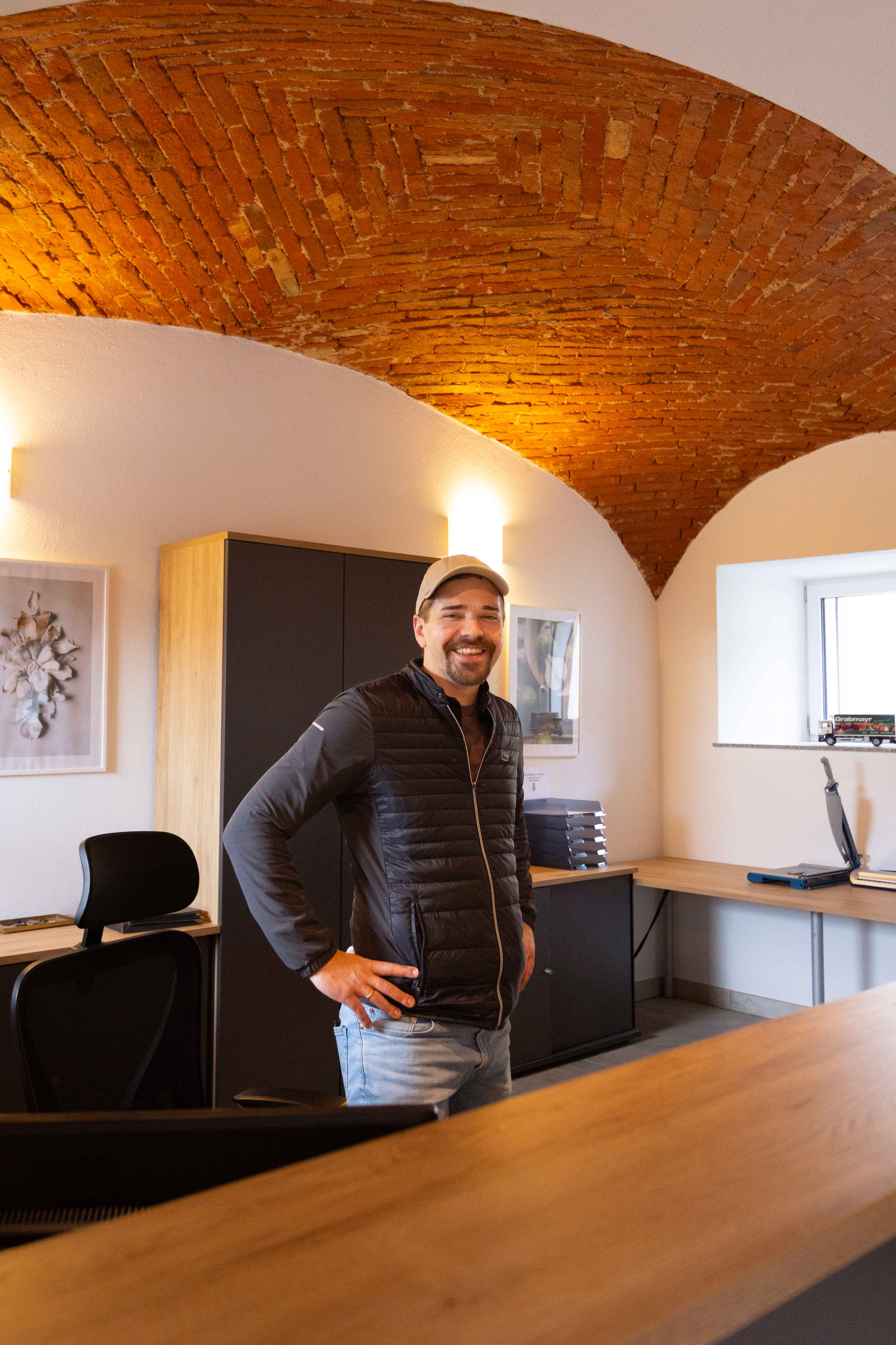 Mann mit Baseballkappe und schwarzer Weste steht lächelnd in einem Büro mit Backsteinbögen an der Decke.