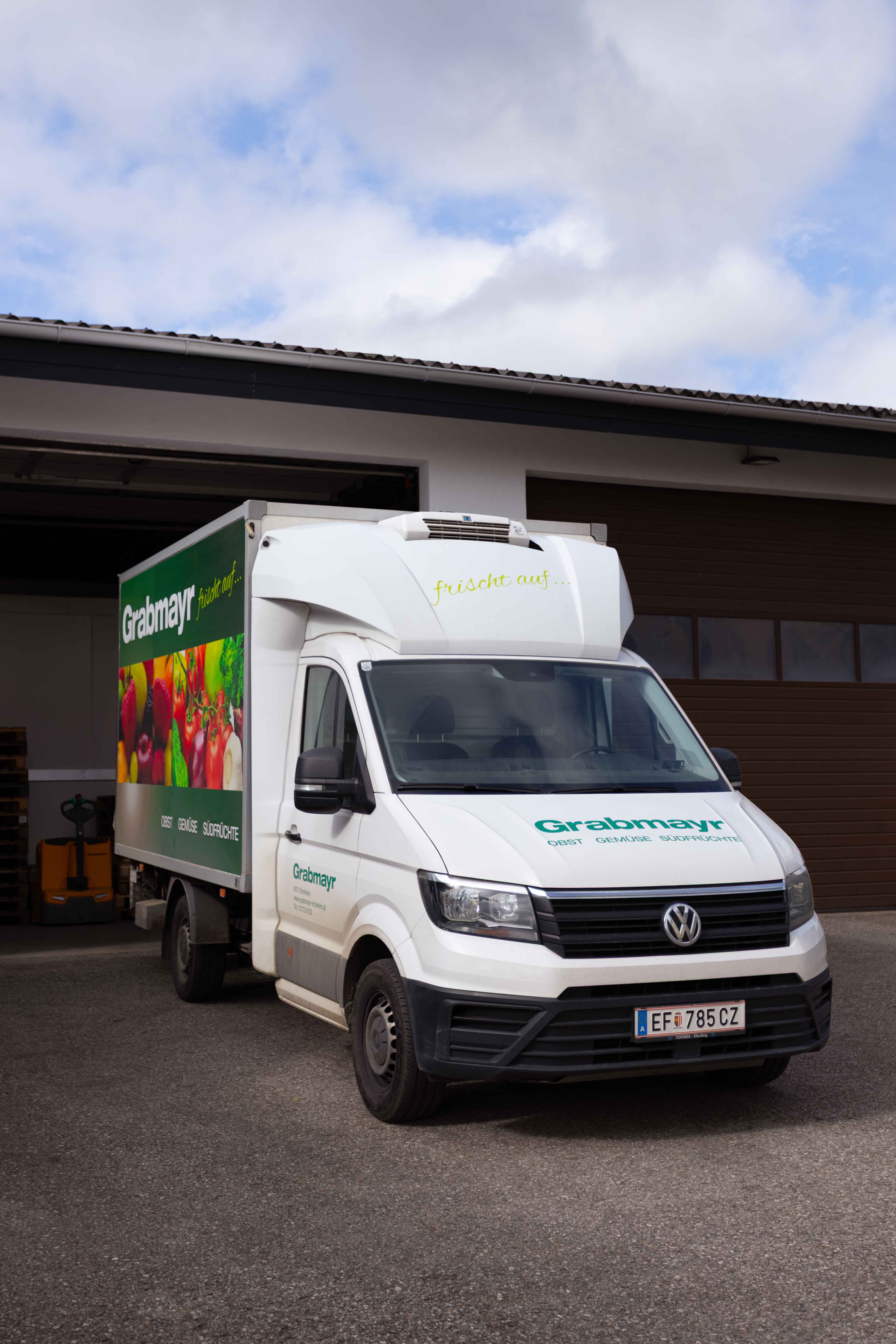 Weißer Lieferwagen mit dem Schriftzug 'Grabmayr' und Bildern von Obst und Gemüse vor einem Lagerhaus.