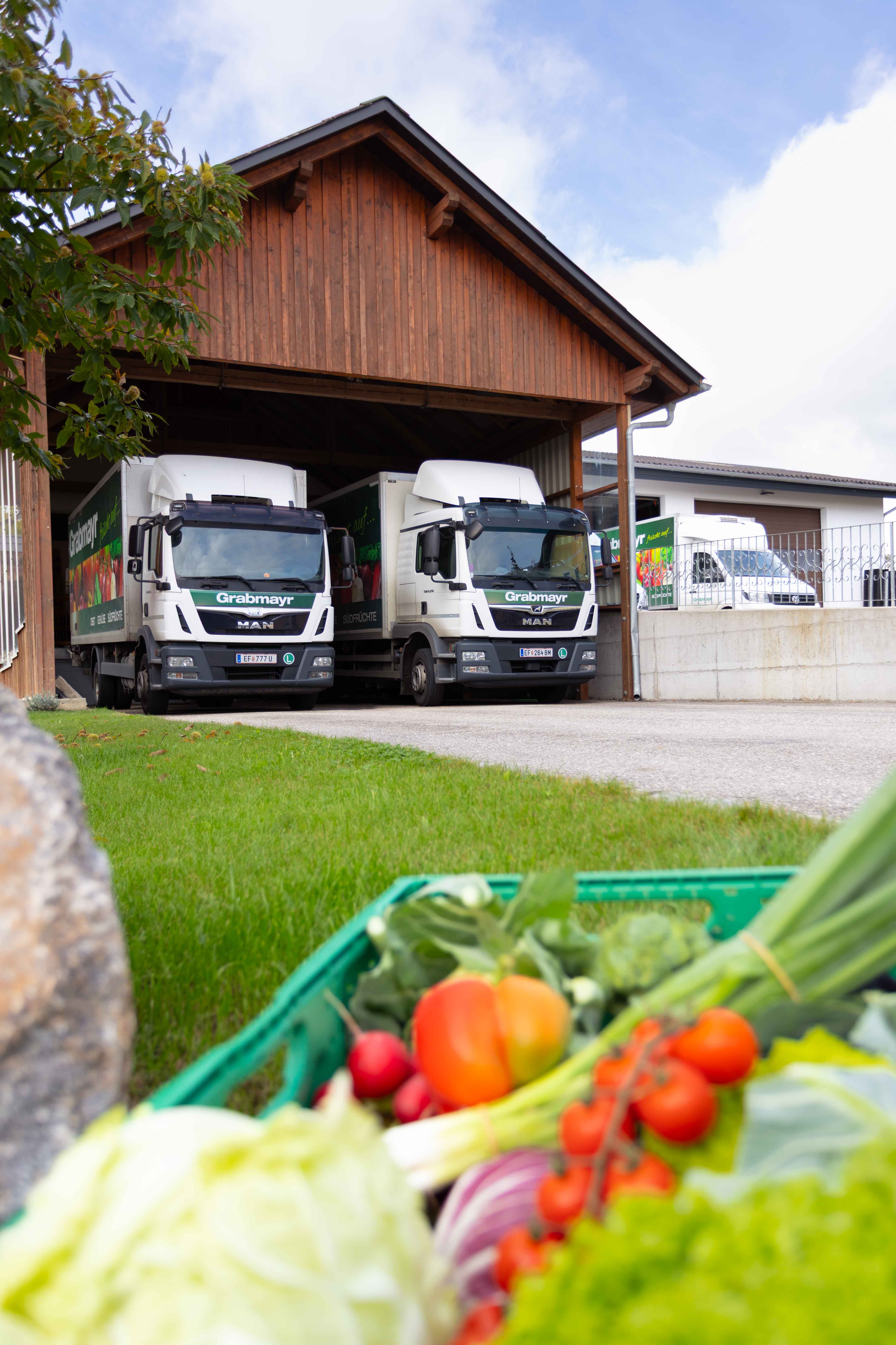 Zwei Grabmayr-LKWs parken in einem Holzschuppen, im Vordergrund eine Kiste mit frischem Gemüse.