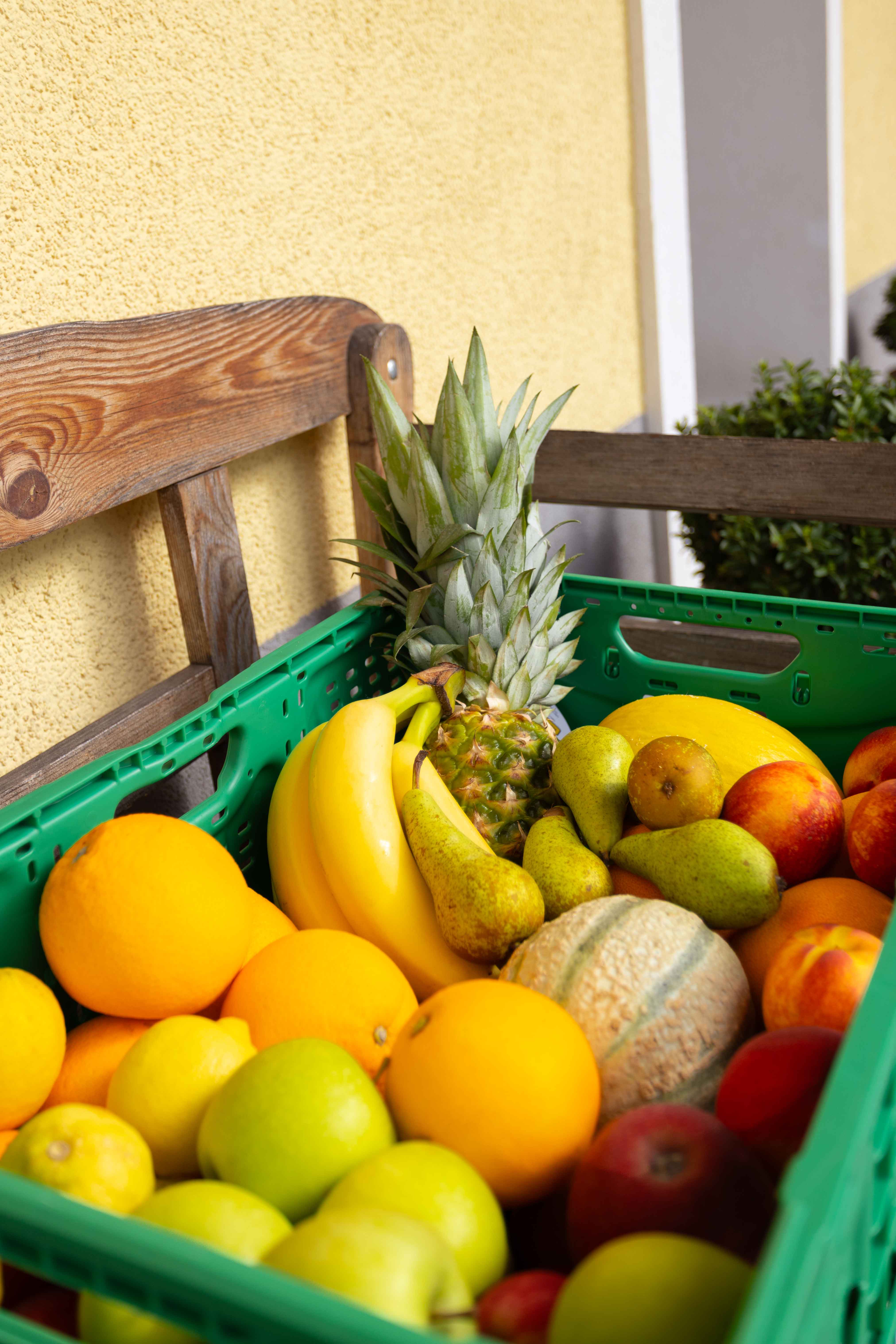 Grüner Kunststoffbehälter gefüllt mit verschiedenen Früchten wie Bananen, Ananas, Birnen, Orangen, Äpfeln und Melone vor einer gelben Wand und einem Holzstuhl.
