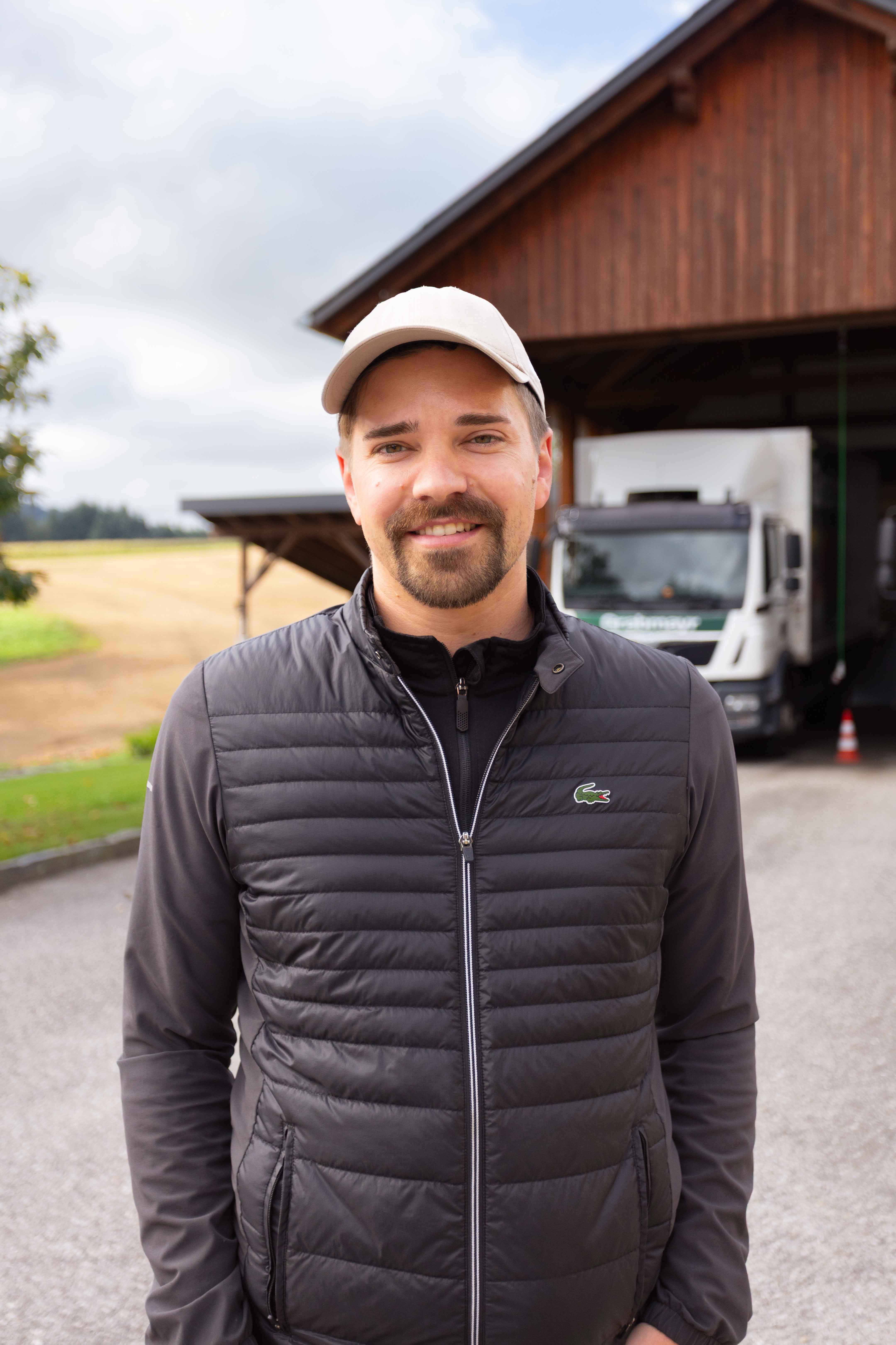 Junger Mann mit Bart, schwarzer Steppweste und heller Kappe steht vor einer Scheune mit einem LKW im Hintergrund.