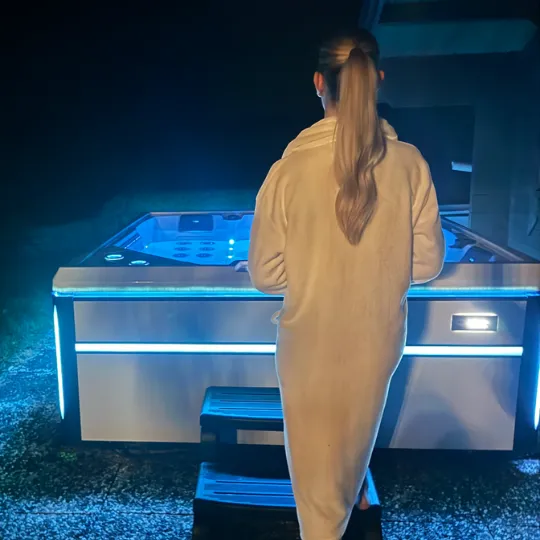 Frau in beiger Robe steht neben blau beleuchtetem Whirlpool im Dunkeln