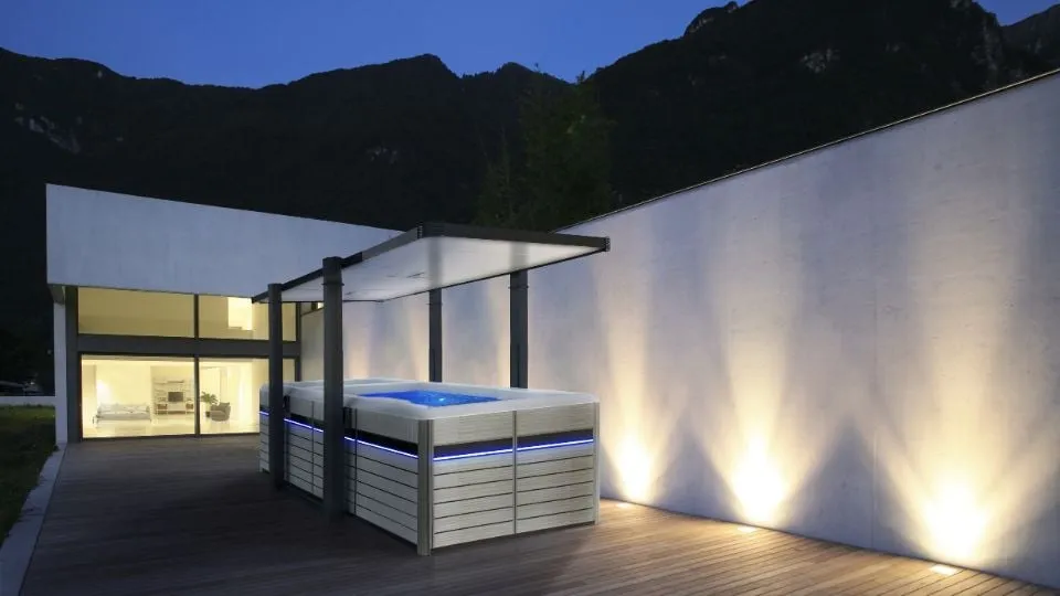 Moderner Whirlpool auf Holzterrasse neben beleuchtetem Haus in Berglandschaft