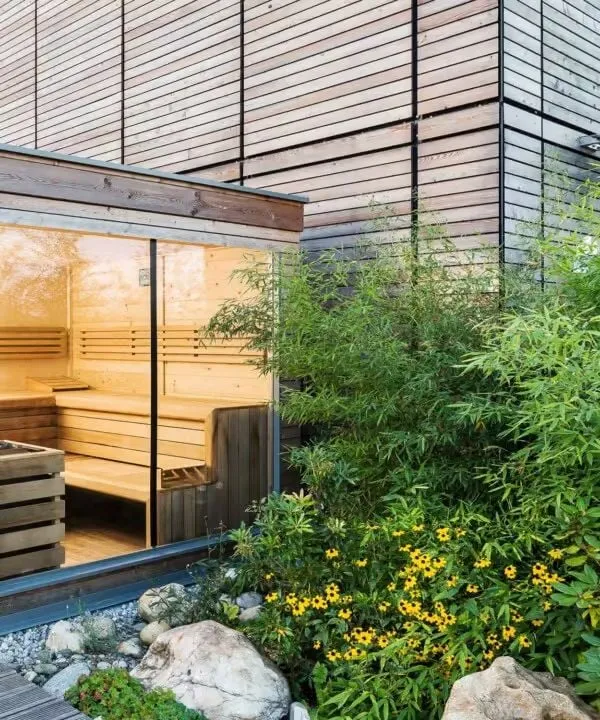 Moderne Sauna mit Glaswand, umgeben von grünen Pflanzen und Steinen