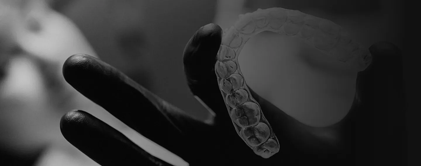 Schwarz-weiß-Aufnahme eines Zahnarztes mit schwarzen Handschuhen, der eine transparente Aligner-Zahnschiene vor einer Patientin hält