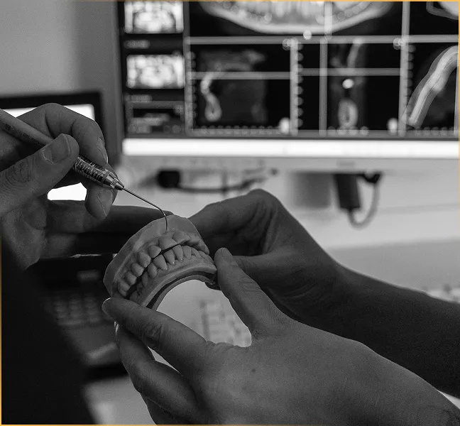 Schwarz-weiß-Aufnahme eines Zahnarztes, der an einem Gebissmodell Behandlungsschritte erklärt, während digitale Röntgen- und 3D-Scans auf dem Monitor sichtbar sind