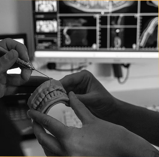 „Schwarz-weiß-Aufnahme eines Zahnarztes, der an einem Gebissmodell Behandlungsschritte erklärt, während digitale Röntgen- und 3D-Scans auf dem Monitor zu sehen sind