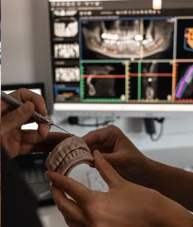 Zahnarzt erklärt an einem Gebissmodell die Behandlungsschritte, während auf dem Monitor digitale Röntgen- und 3D-Scans sichtbar sind