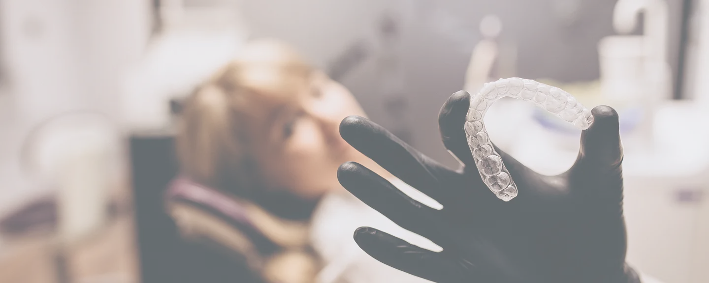 Zahnarzt in schwarzen Handschuhen hält eine transparente Aligner-Zahnschiene vor einer Patientin im Behandlungsstuhl