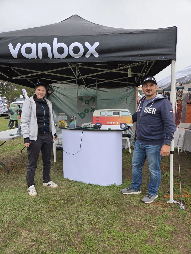 Two people standing at a Vanbox booth under a black canopy tent with promotional materials on the table.