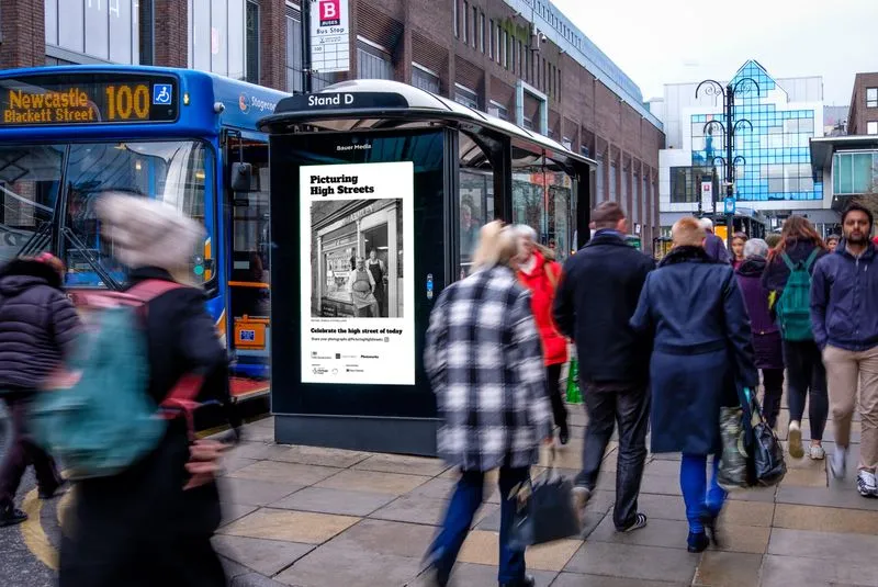 Digital bus shelter screen in Newcastle next to a static bus and a lot of people walking