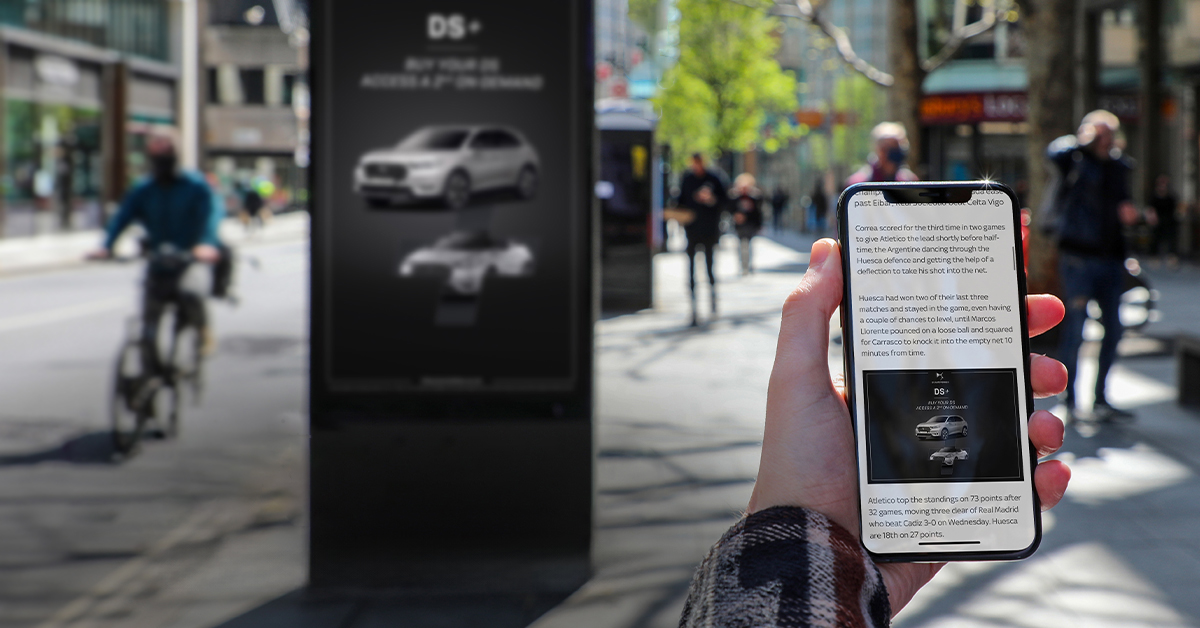Person holding up their phone in front of a digital advertising screen which is showing the same add on their phone as is showing on the digital screen