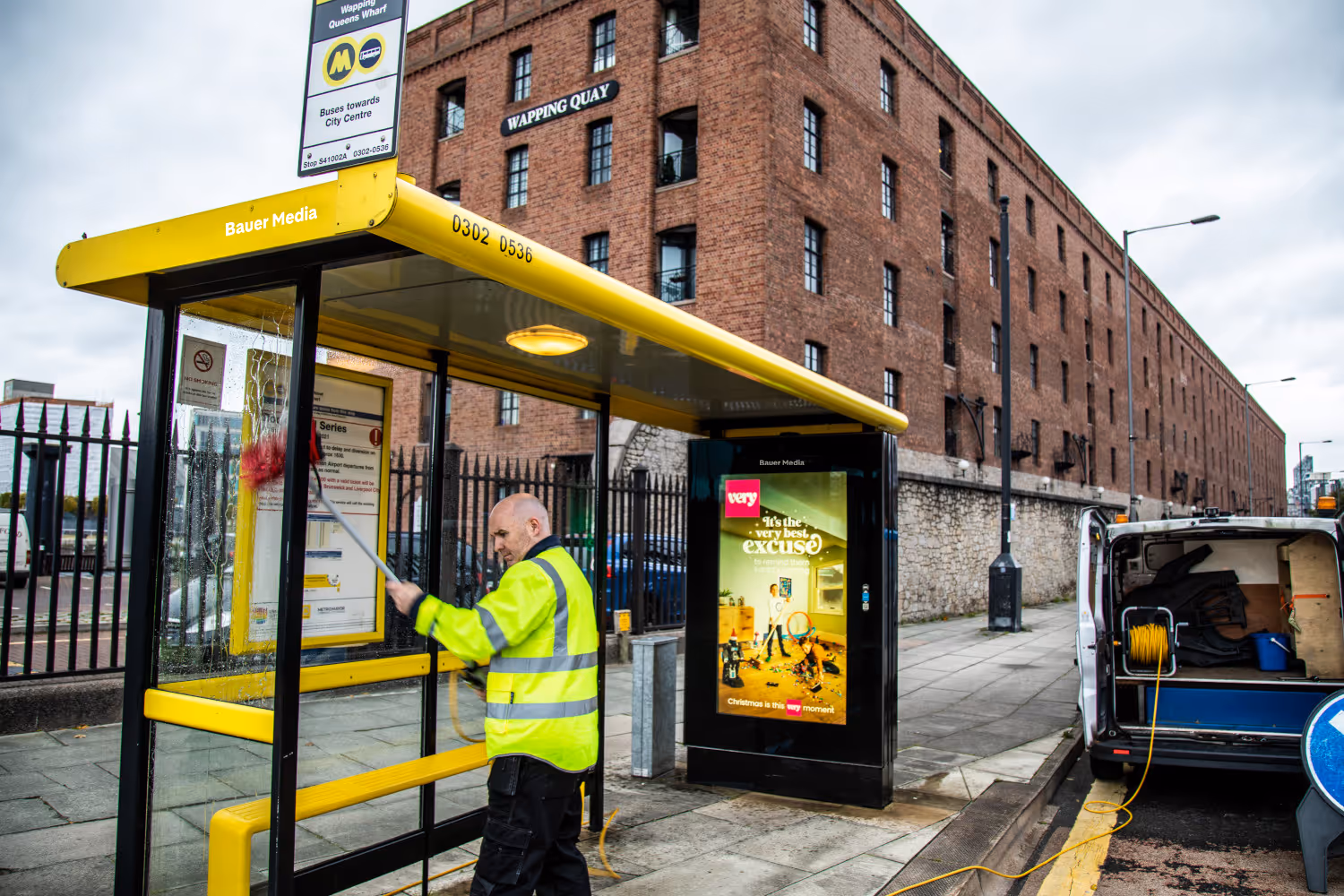 A male Bauer Media Outdoor construction worker cleaning a yellow framed bus shelter in front of a red brick building