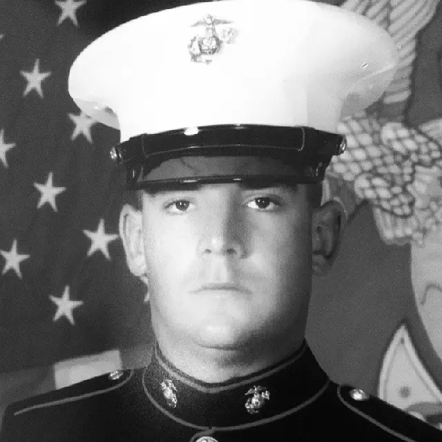 Black and white portrait of a young male Marine in uniform with a white cap, standing in front of an American flag.