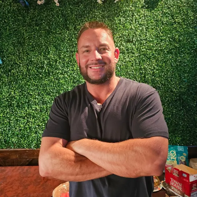 Smiling man with a beard and short hair wearing a black t-shirt, standing with arms crossed in front of a green leafy background.