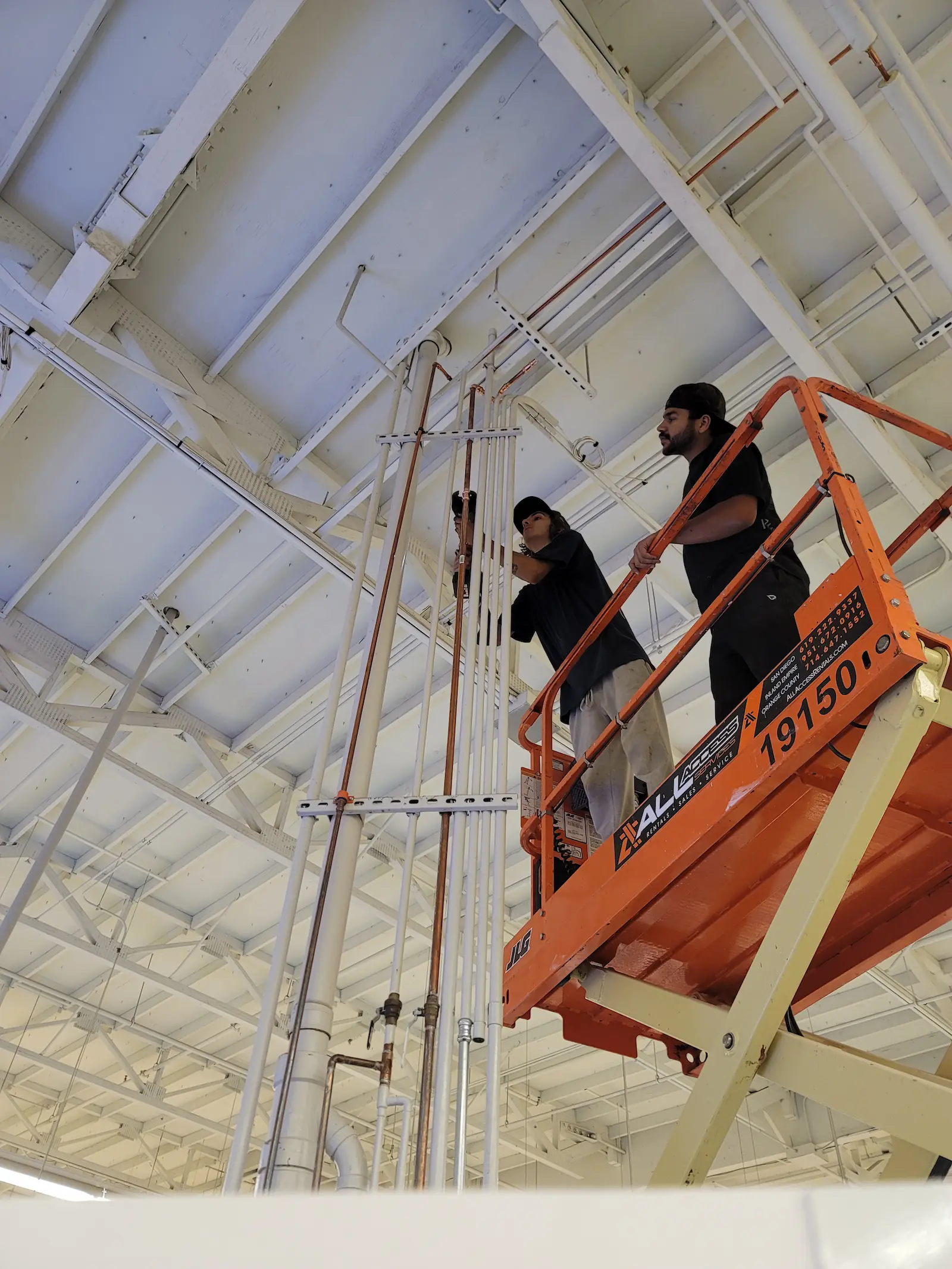 Two men working on pipes on an elevated orange lift inside a spacious industrial building with a white ceiling.