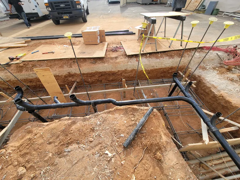 Construction site trench with black plumbing pipes and metal rebar framework, surrounded by dirt piles and caution tape.