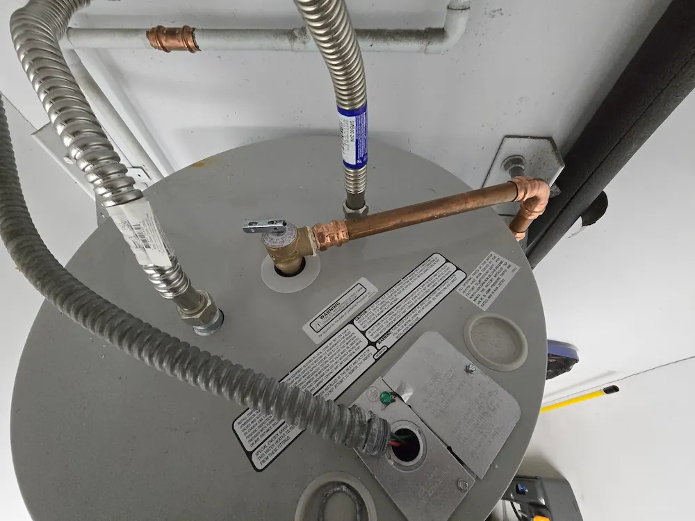 Top view of a gray water heater with copper piping and flexible metal conduit connections.