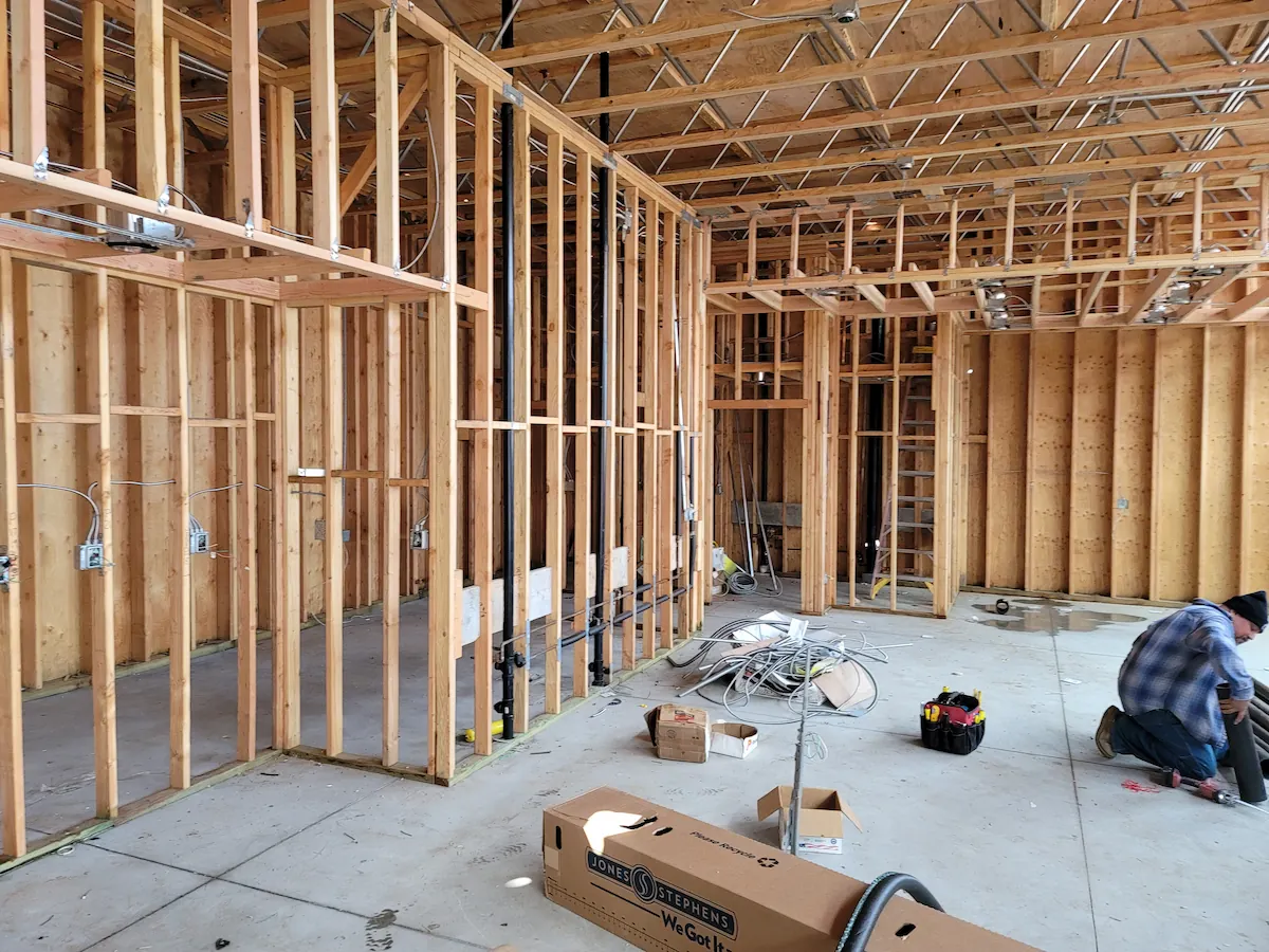Interior of a building under construction showing wooden framing, electrical wiring, and a worker kneeling on the floor.