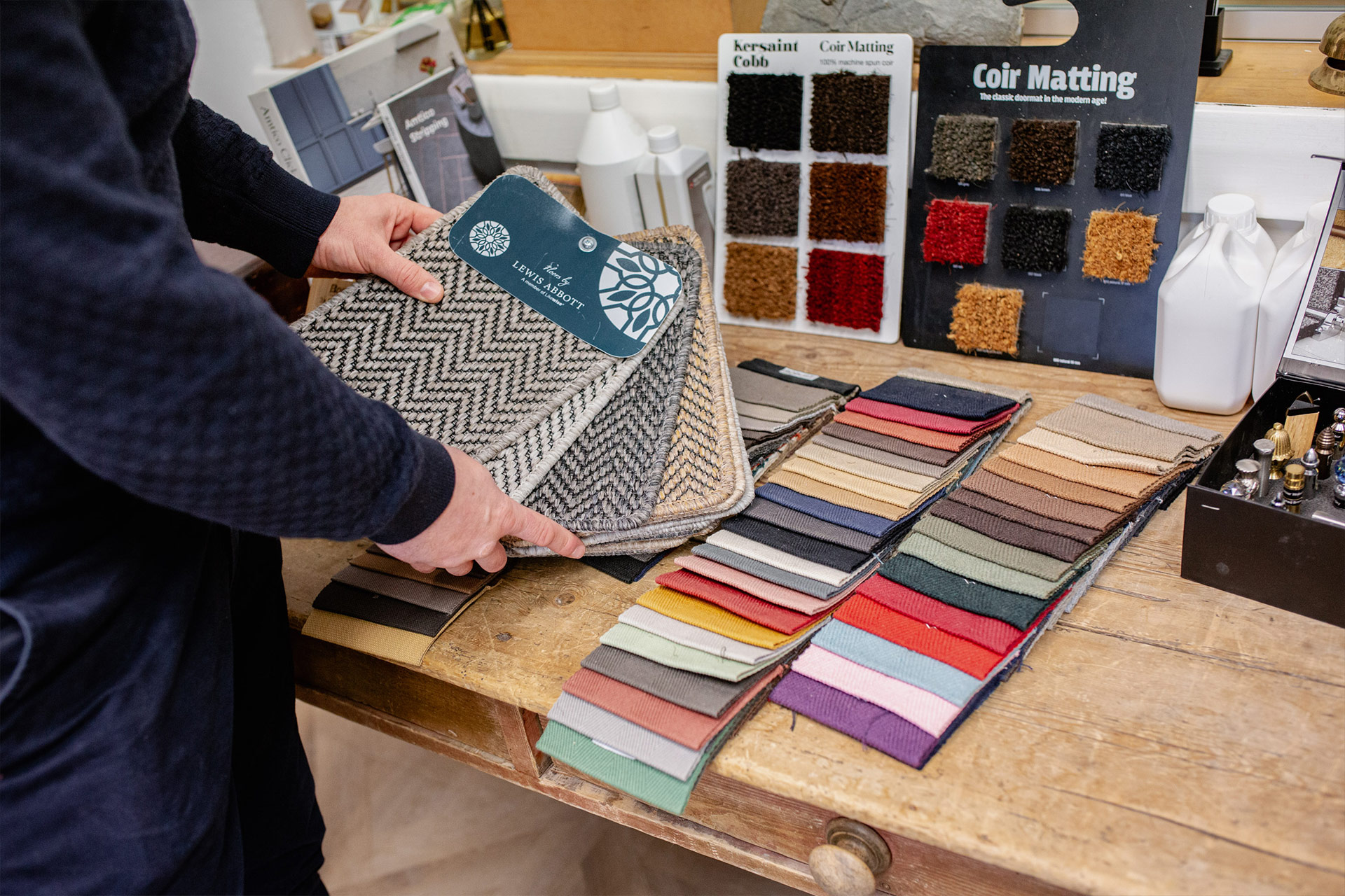 Person holding carpet samples over a wooden table displaying fabric swatches and coir matting color cards.