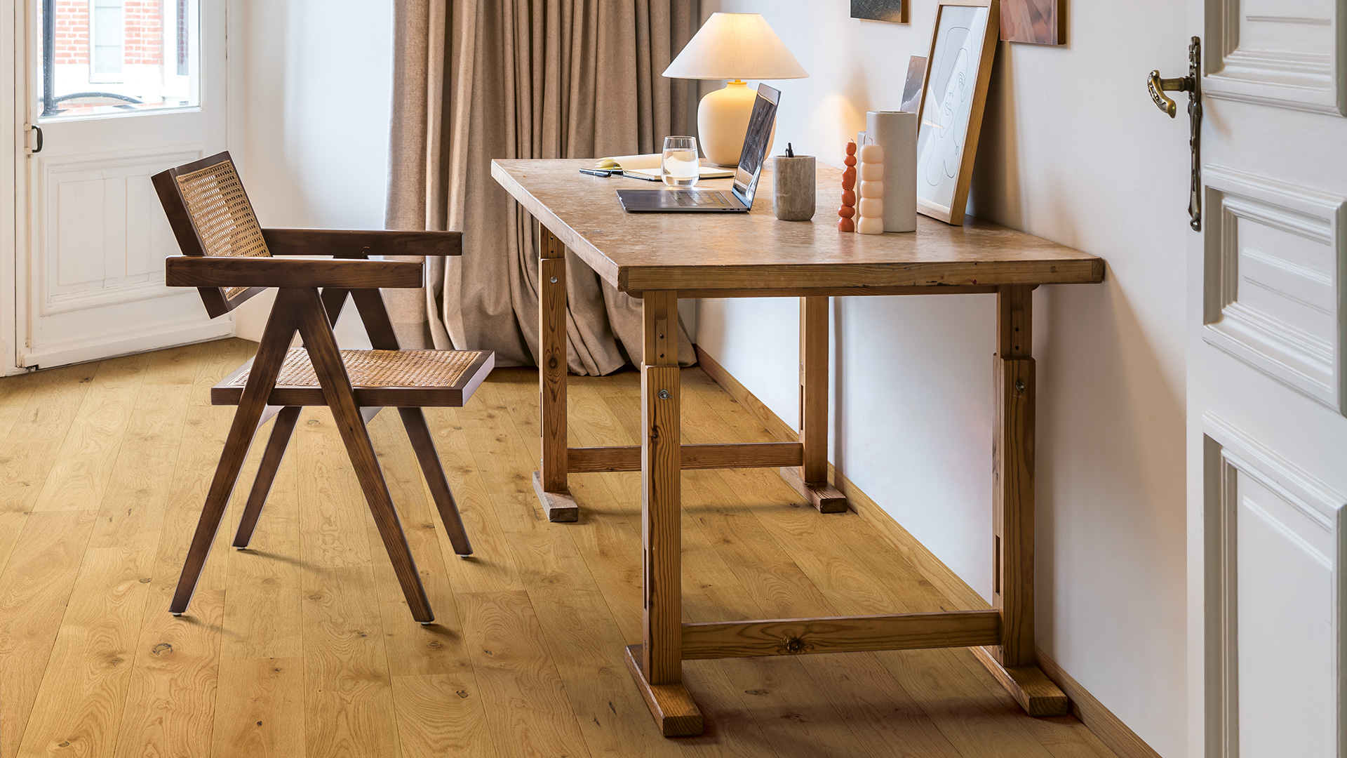 Quick-Step Wood Effect Laminate Flooring Minimalist wooden desk with laptop, lamp, glass of water, and decorative items next to a woven wooden chair on hardwood floor.