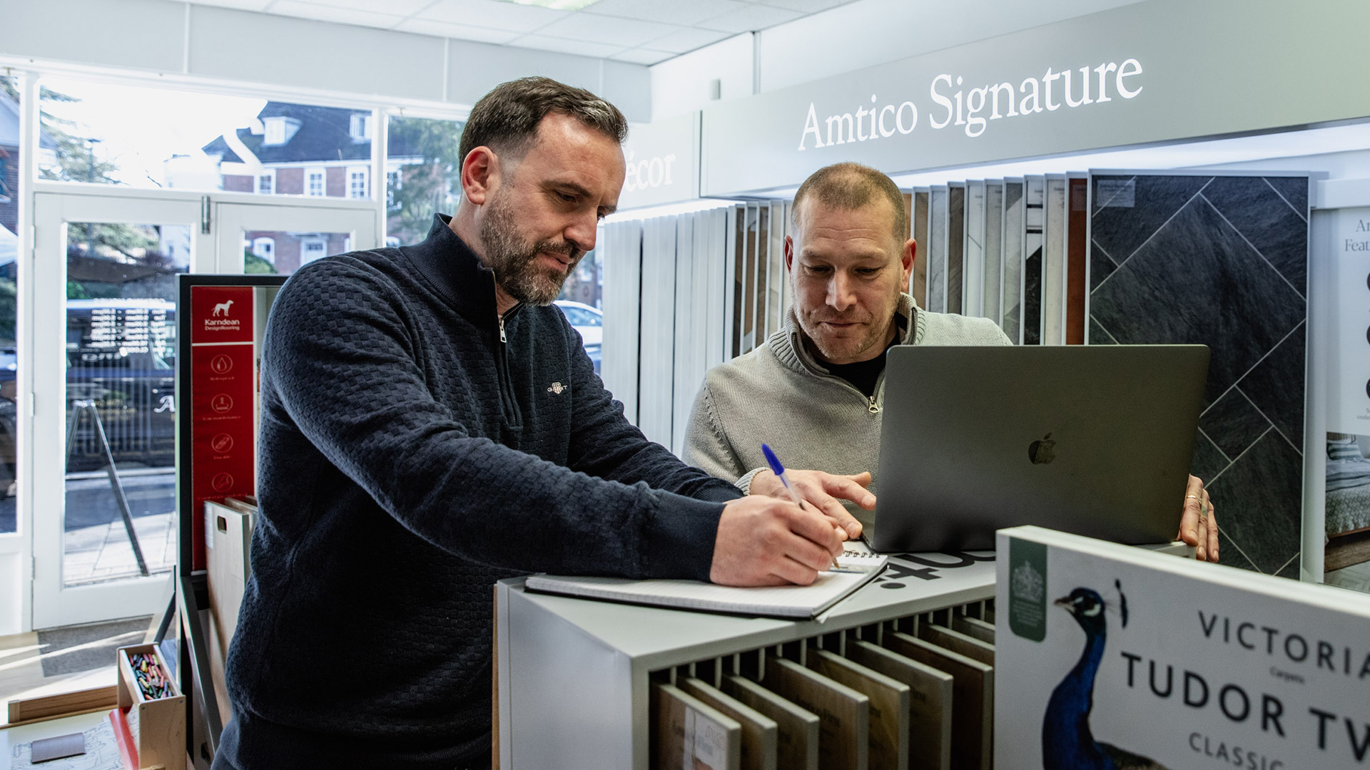 Two men at a showroom counter at Ammonite Flooring reviewing flooring samples, one writing in a notebook and the other using a laptop.