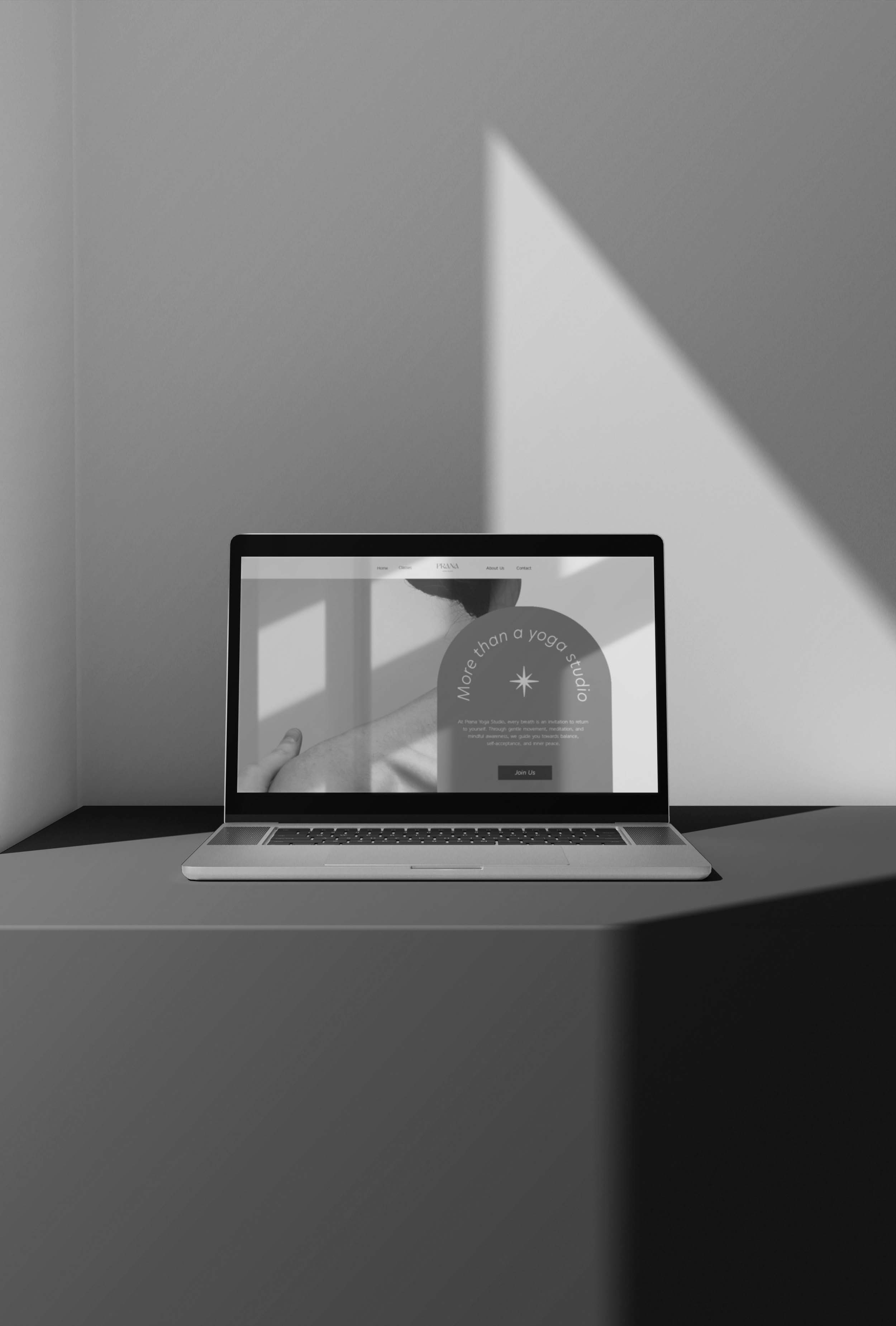 A laptop sits on a dark surface in a minimalist room with light and shadow patterns on the wall. The screen displays a website, though the text is mostly unreadable. The scene is in black and white.