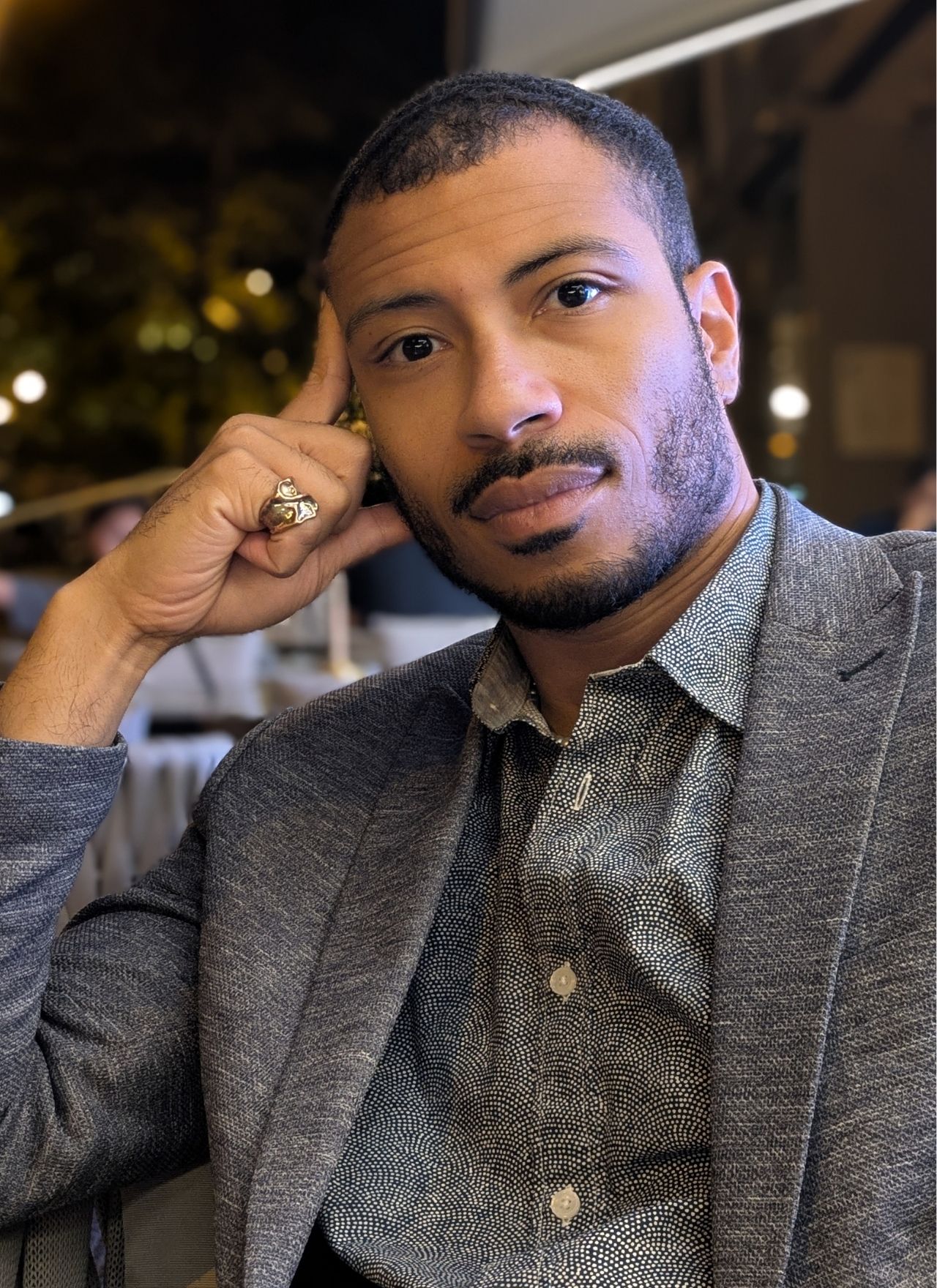 Man with short hair and beard wearing a patterned shirt and gray blazer, resting his head on his hand and looking at the camera.