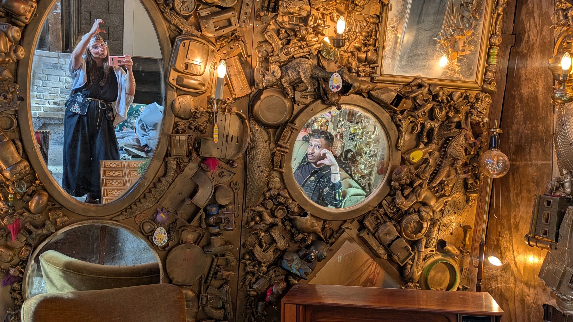 Decorative wall with three mirrors surrounded by vintage mechanical parts and two people reflected, one standing and one seated.