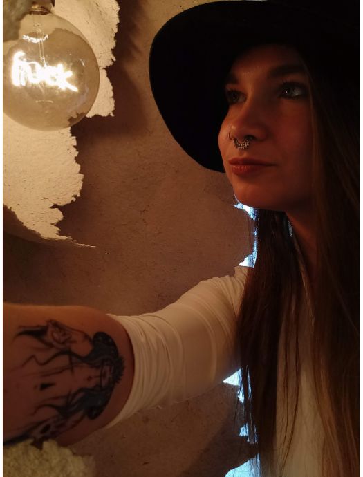Woman with long hair, wearing a black hat and white shirt, looking at a glowing light bulb, showing a tattoo on her forearm.