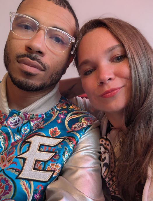 Close-up selfie of a man wearing clear glasses and a colorful floral jacket with the letter E, alongside a woman with long brown hair and a nose ring.