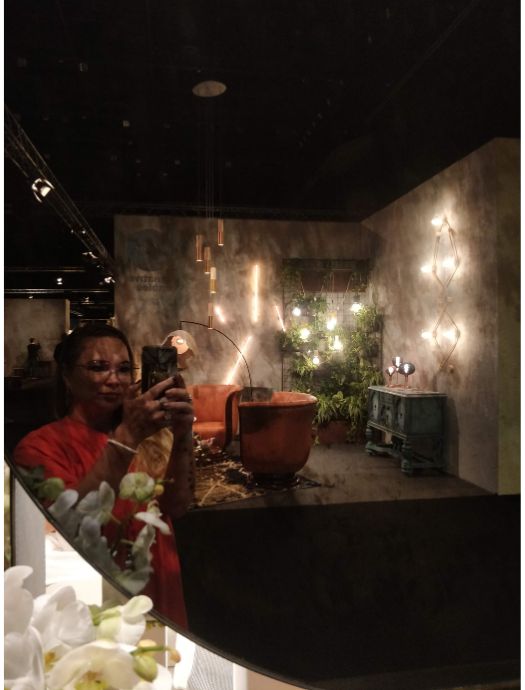Reflection of a person taking a photo with a smartphone, showing a warmly lit room with orange chairs, green plants, and wall-mounted light fixtures.