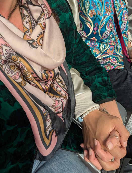 Two people holding hands, one wearing a floral patterned blue and pink top and the other wearing a dark green textured jacket with a decorative scarf.