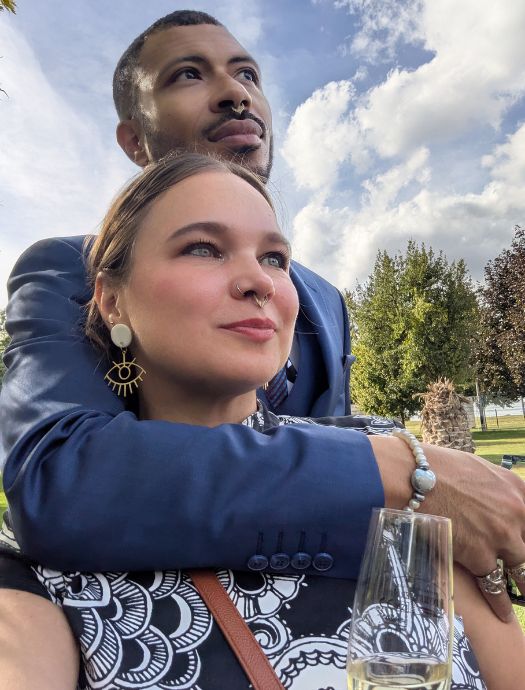 Couple embracing outdoors with a scenic sky and trees in the background, woman holding a glass of white wine.