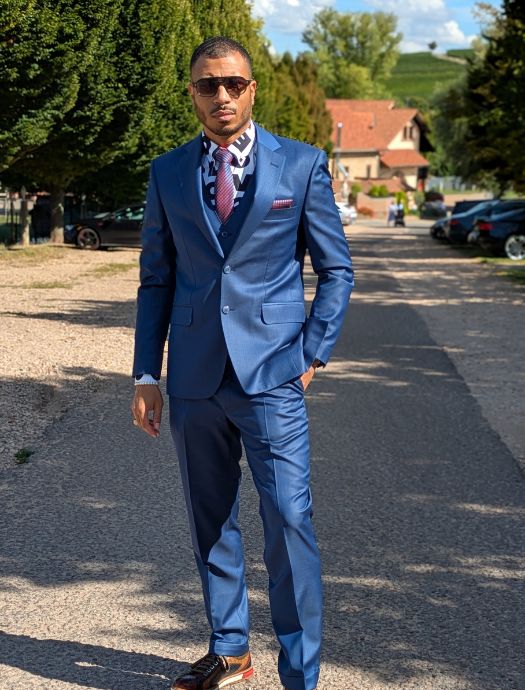 Man in a blue suit with a patterned shirt, red tie, and sunglasses standing on a sunny paved street with greenery and houses in the background.