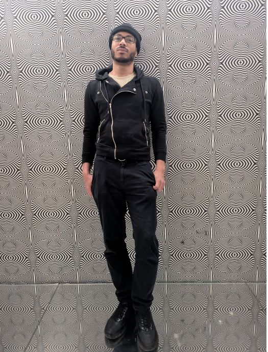 Man wearing glasses, a black beanie, and black clothes standing against a black and white wavy patterned wall.