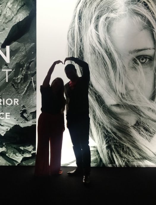 Silhouettes of two people forming a heart shape with their arms in front of a large black-and-white photo of a woman’s face with flowing hair.