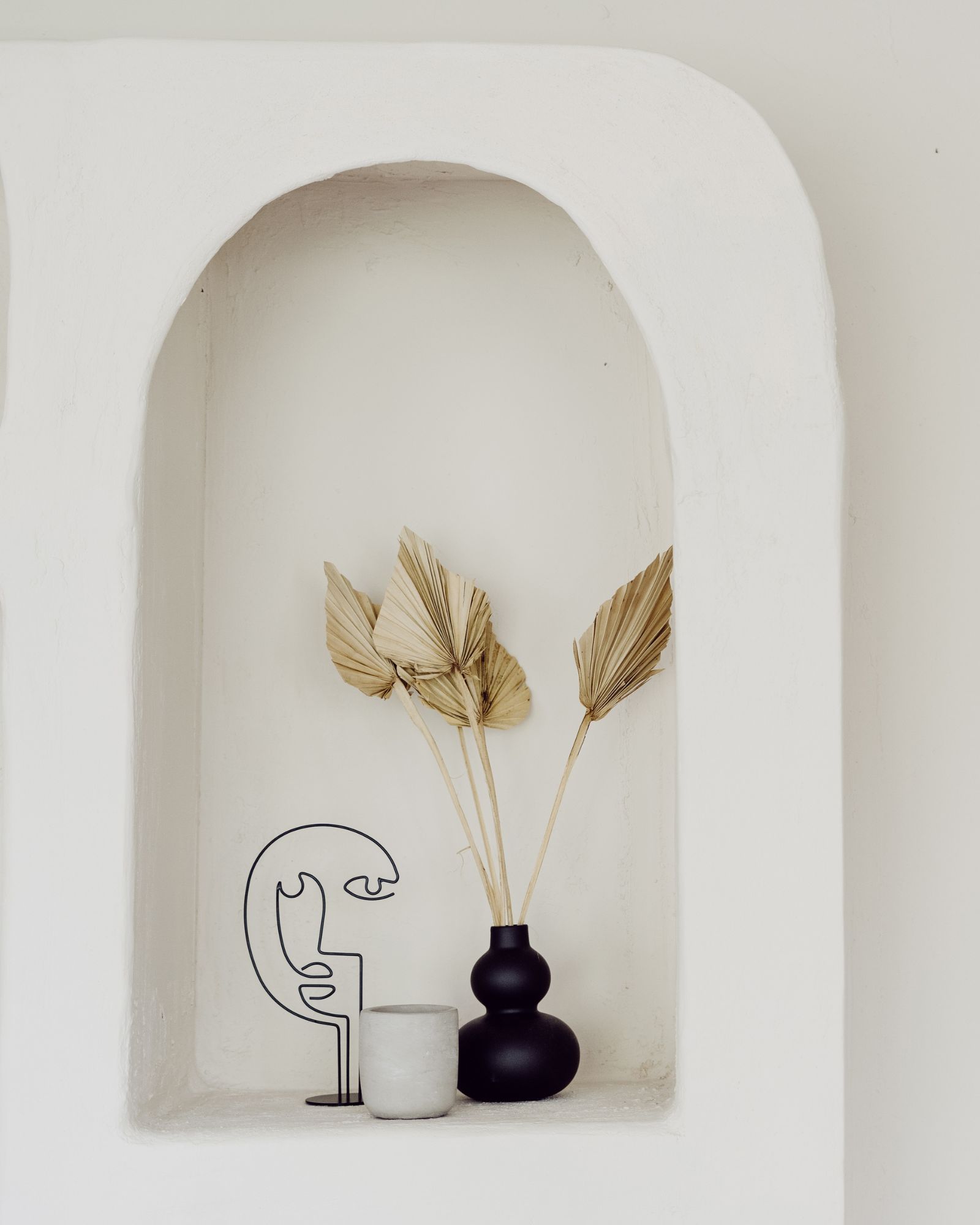 White alcove shelf with a black vase holding dried palm leaves, a gray stone cup, and a black wire face sculpture.